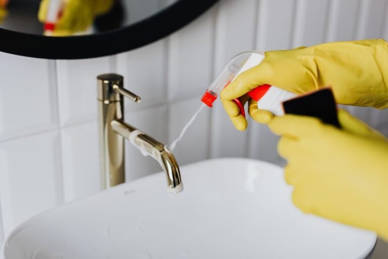Person wearing yellow rubber gloves spraying cleaner on a bathroom sink faucet