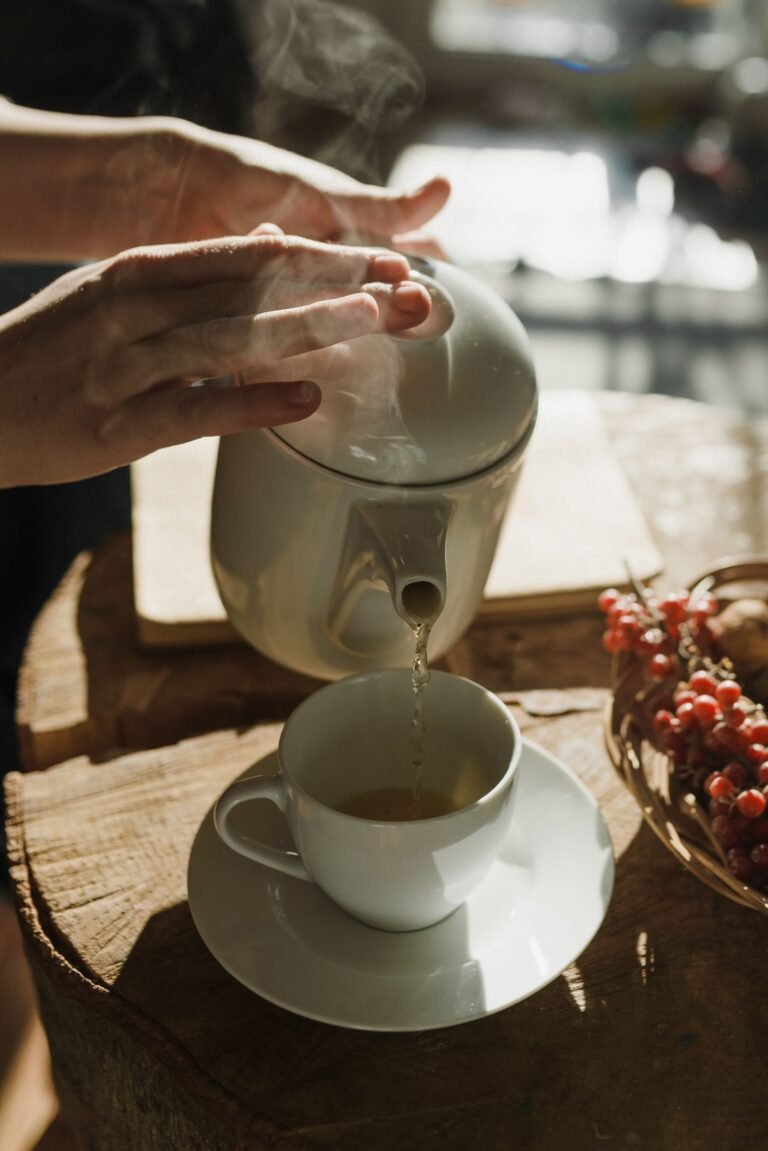 Warm tea being poured from a white teapot into a cup in a sunlit kitchen