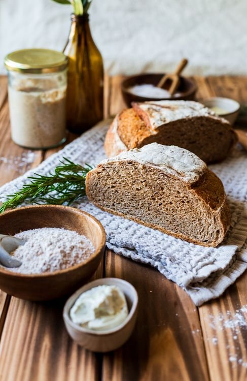 Sourdough bread on a rustic table with a jar of starter and a wooden bowl of flour
