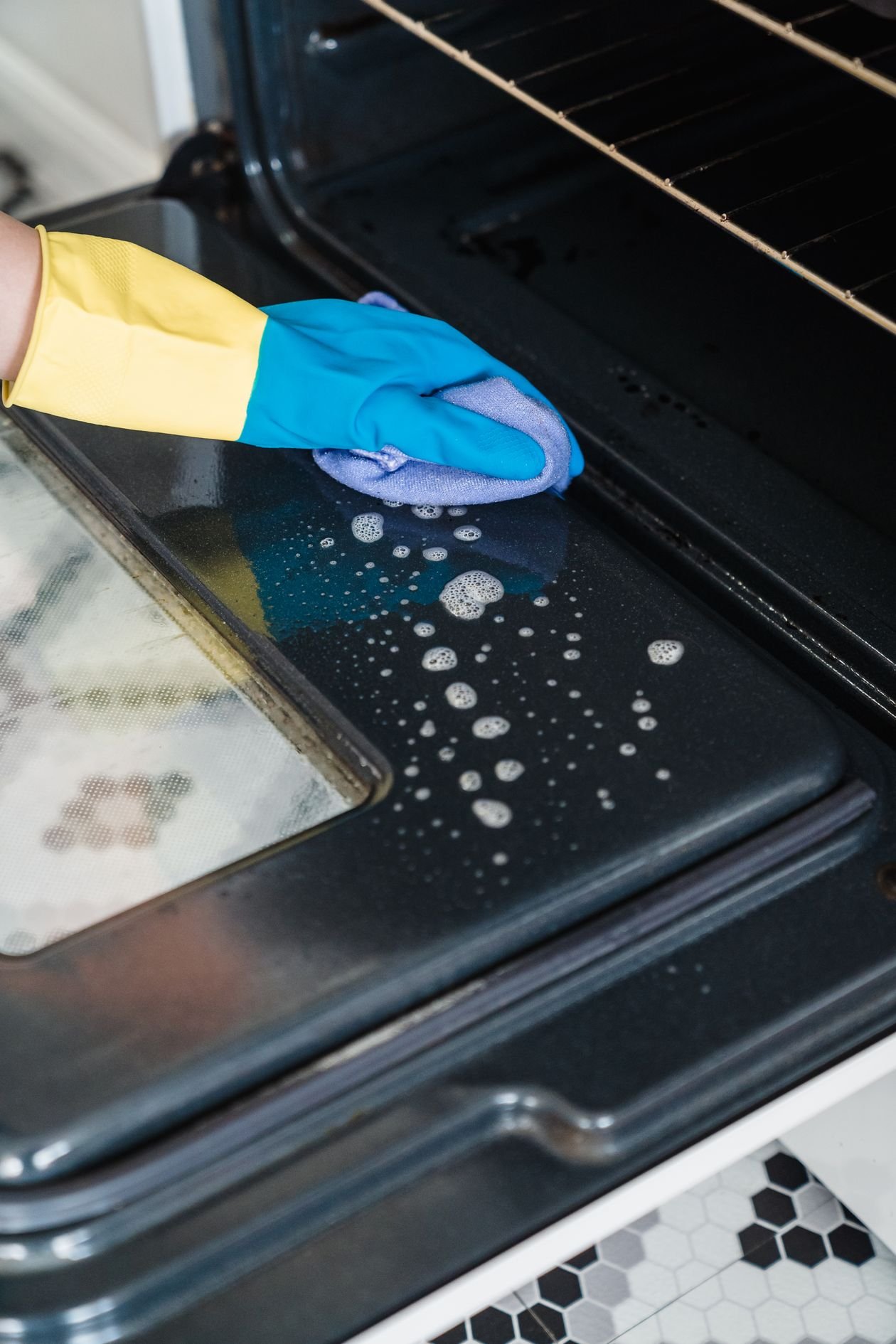 Close up of a gloved hand cleaning an oven door with spray foam and cloth